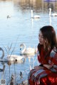 A woman sitting on the shore of a lake with swans in the background.