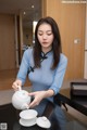 A woman in a blue dress pouring tea into a white teapot.